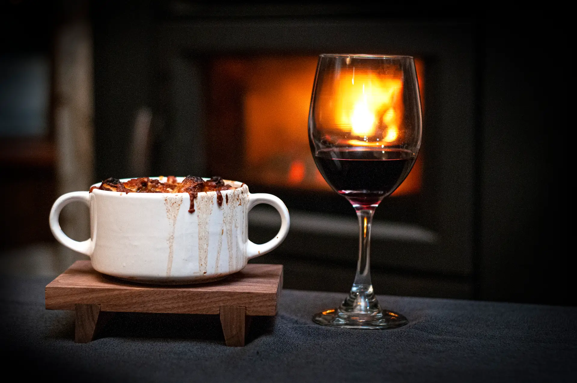 Bol blanc à deux anses rempli de soupe à l’oignon gratinée qui déborde, posé sur un socle de bois à côté d’un verre de vin rouge, devant un foyer allumé